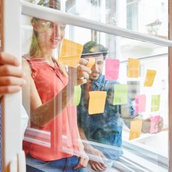 Team notiert kreative Ideen auf bunten Zetteln auf einer Transparenten Scheibe im Start-Up Workshop