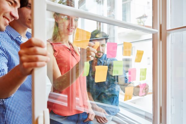 Team notiert kreative Ideen auf bunten Zetteln auf einer Transparenten Scheibe im Start-Up Workshop